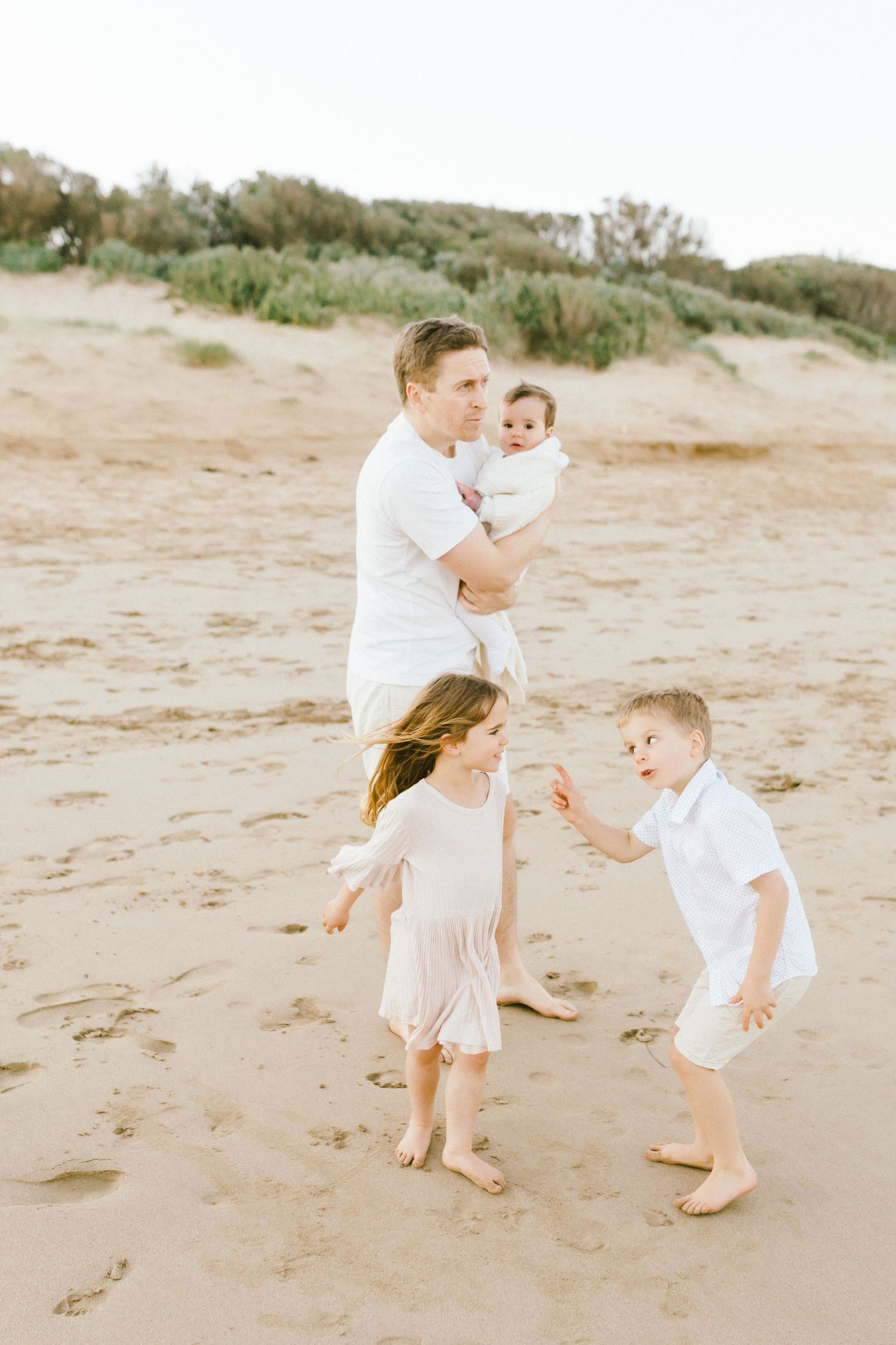 A family in white clothing walks and plays together on a sandy beach during a sunny day.