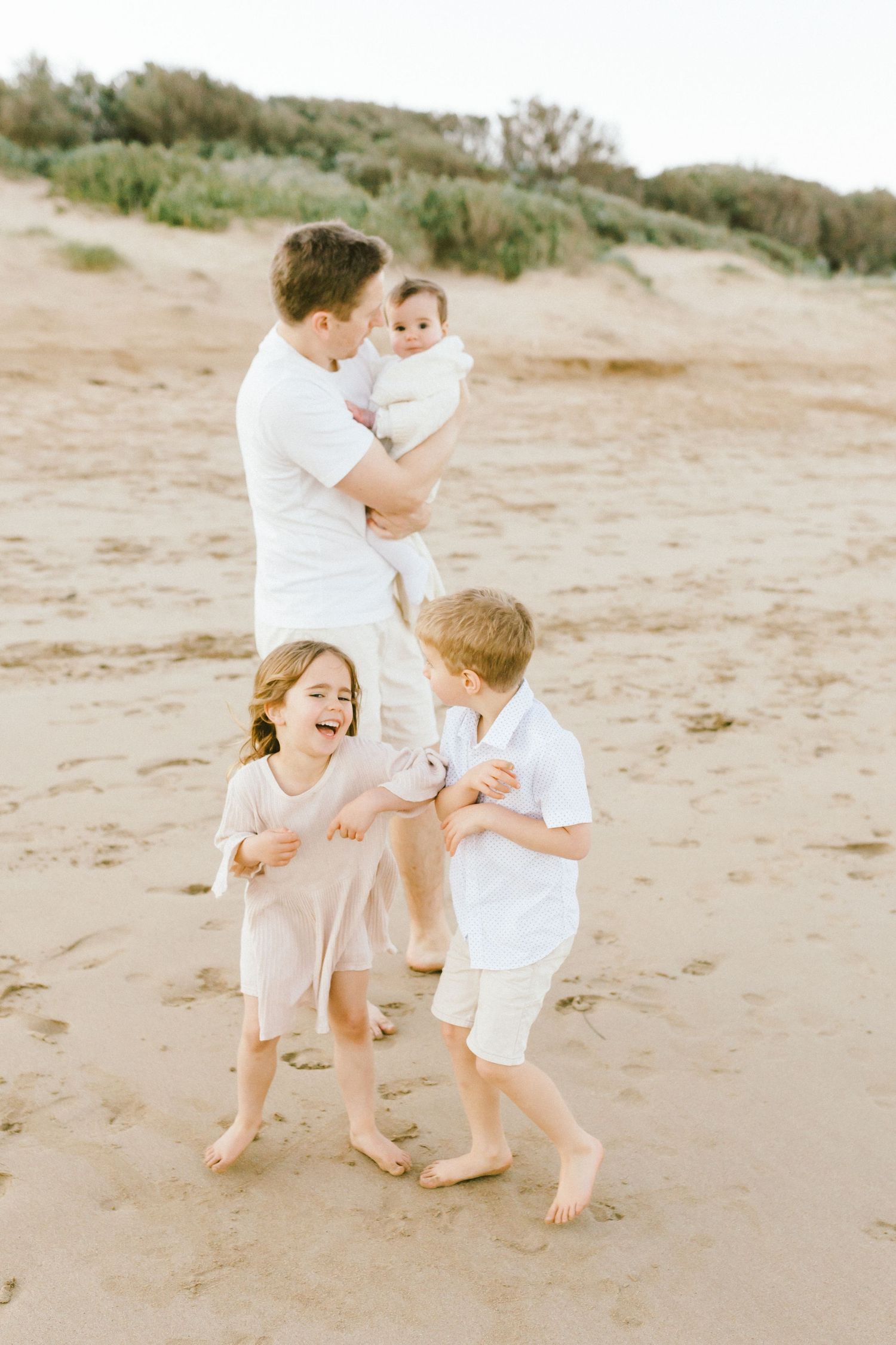 A family in white clothing plays and laughs together on a sandy beach during a bright sunny day.