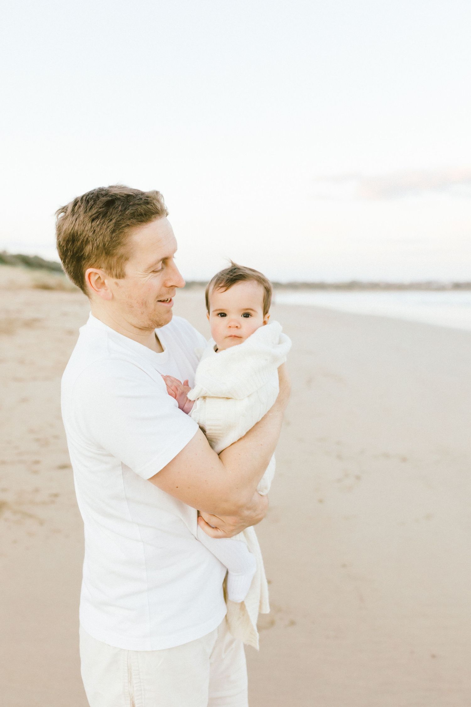 Parent and baby share tender moments together on a serene beach with soft lighting and white attire.