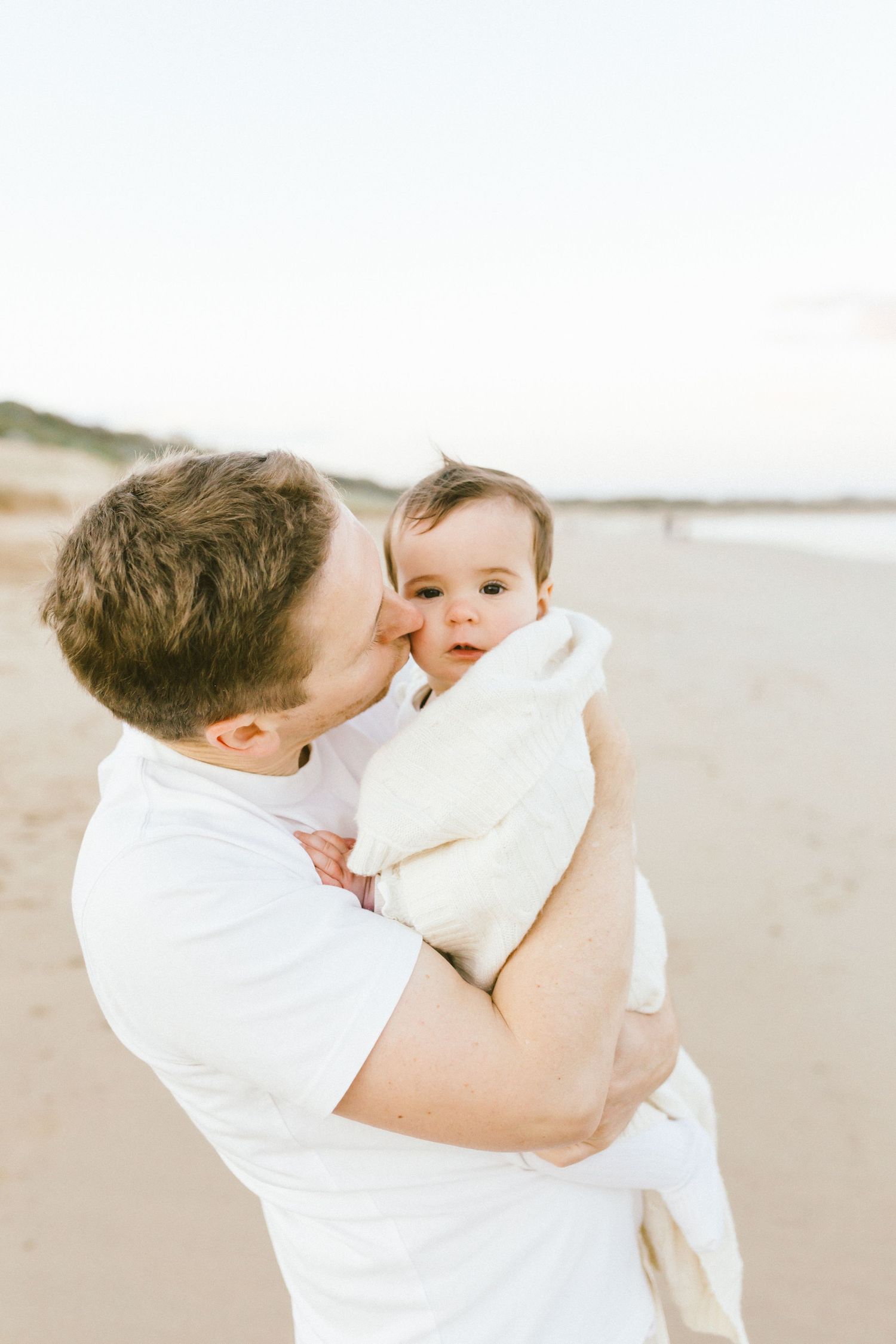 Parent in white shirt holds and kisses baby wearing white on sunny beach during tender family moment.