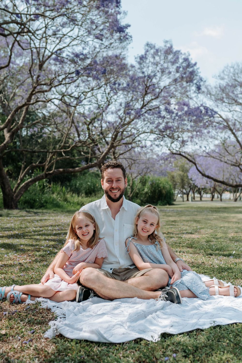 A family relaxes together on a white blanket under blooming jacaranda trees in a grassy park setting.