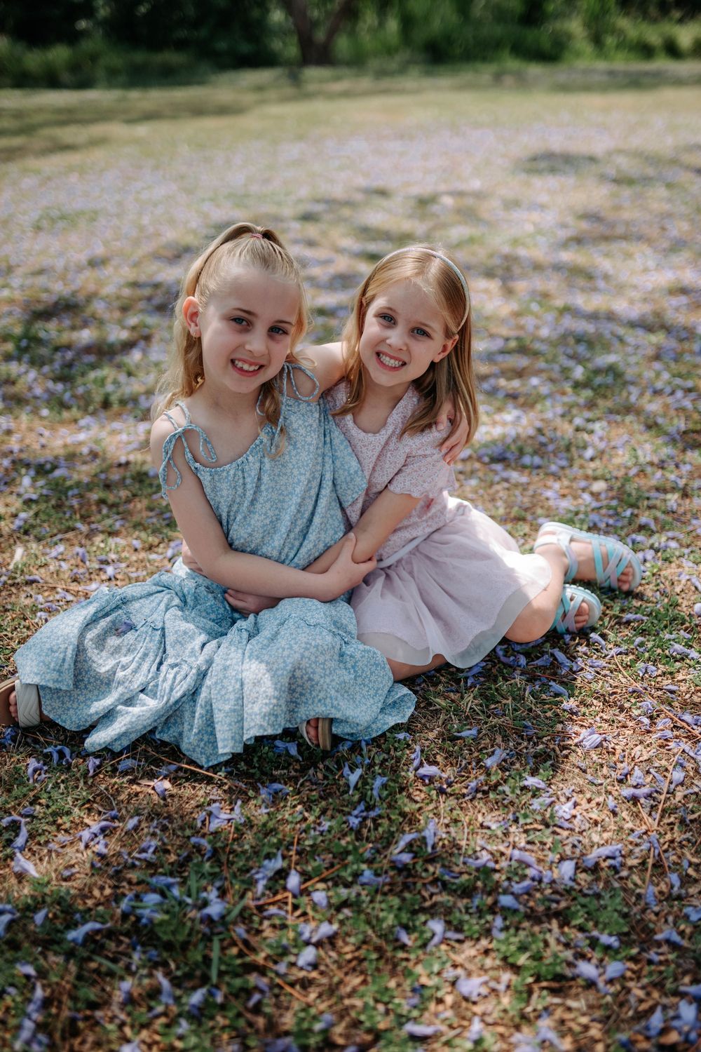 Two young children in blue and pink dresses laugh together while sitting in a field of scattered flower petals.