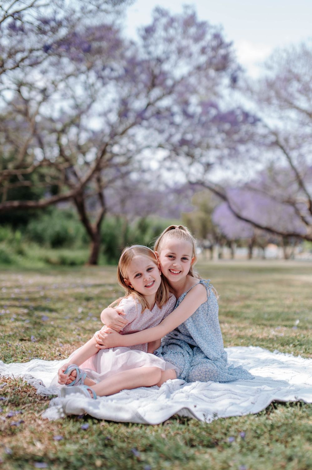 Two young children laying on a white blanket under blooming cherry blossom trees in a spring garden setting.