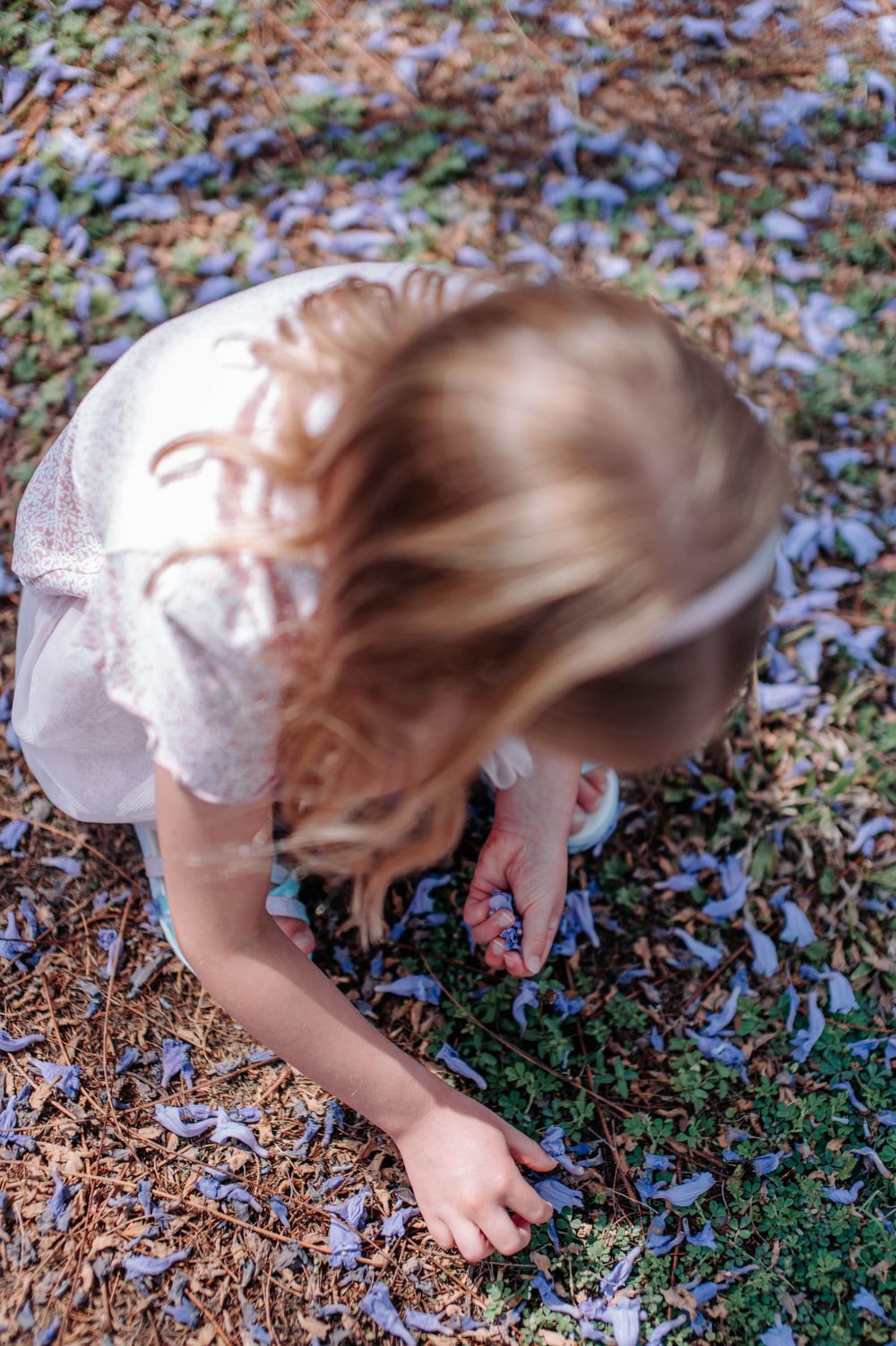 A child in a white dress bends down to explore colorful flowers scattered on the ground.