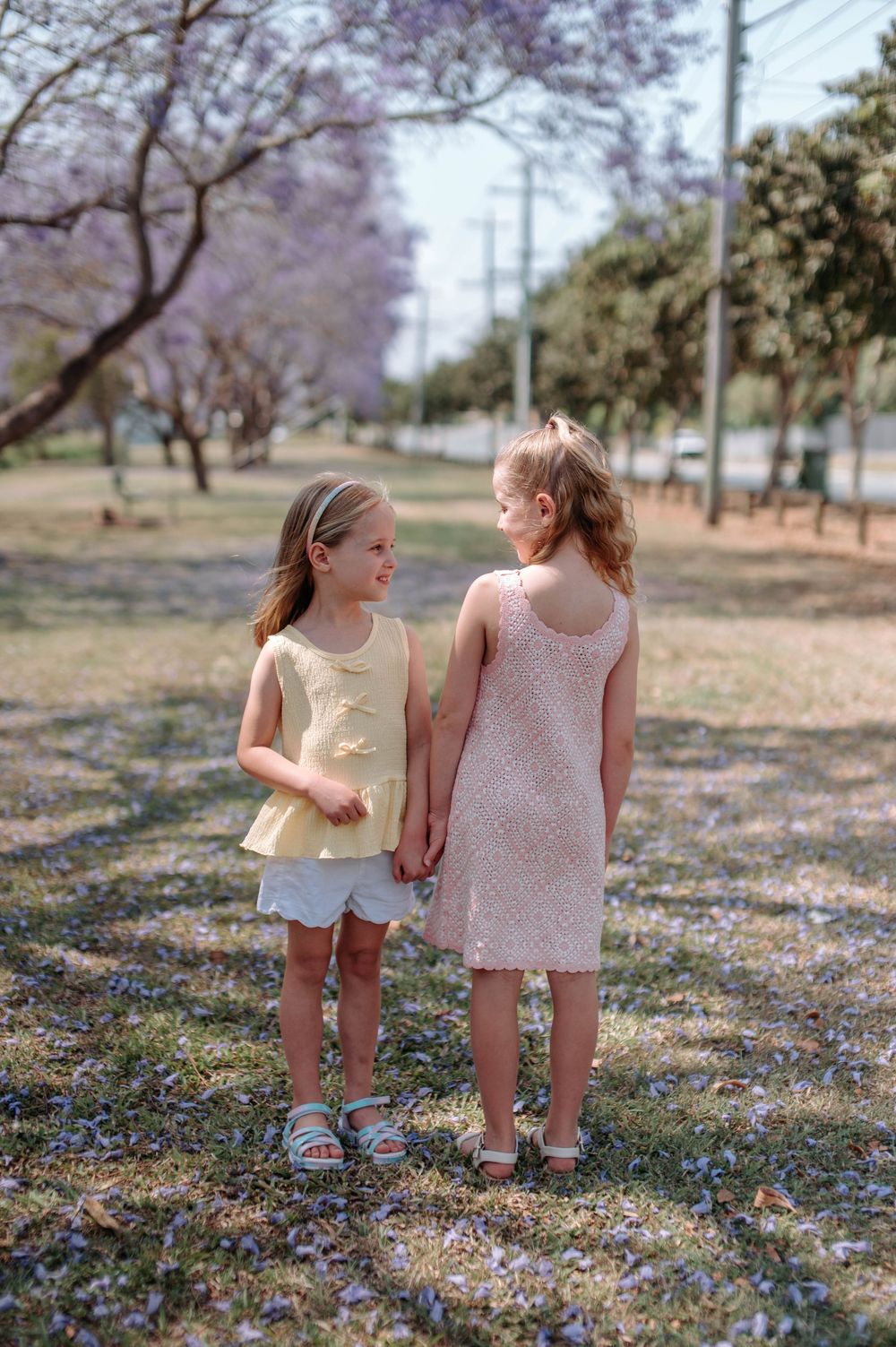 Two young children in light colored dresses walk together through a field of scattered purple flowers.