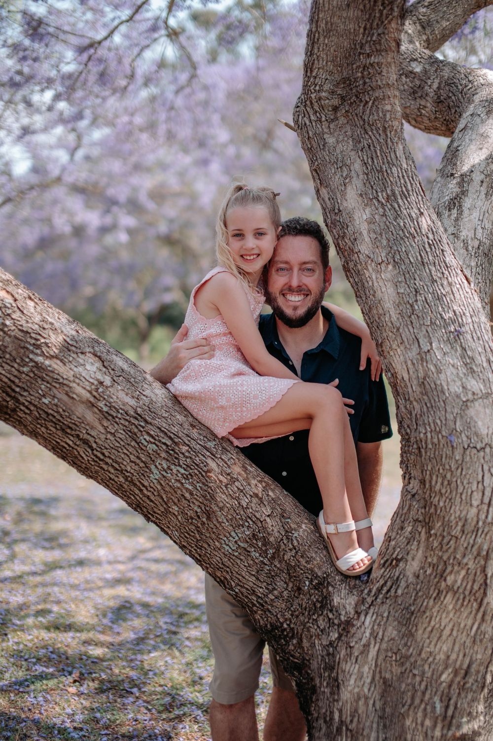 A playful family moment captured under a flowering jacaranda tree with purple blossoms scattered on the ground.