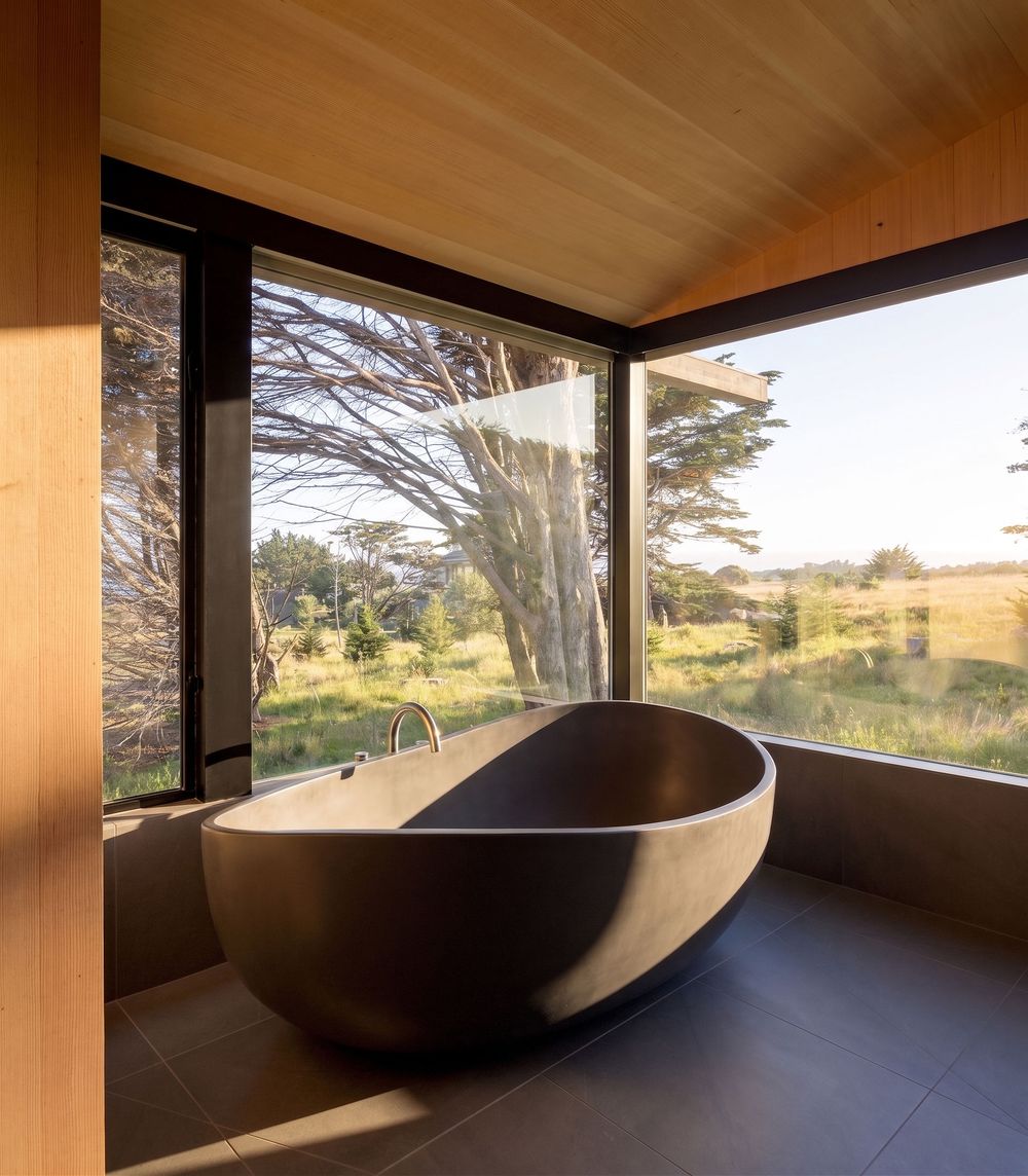 Freestanding dark stone bathtub beside floor-to-ceiling windows overlooking lush green landscape in a modern wood-paneled bathroom.