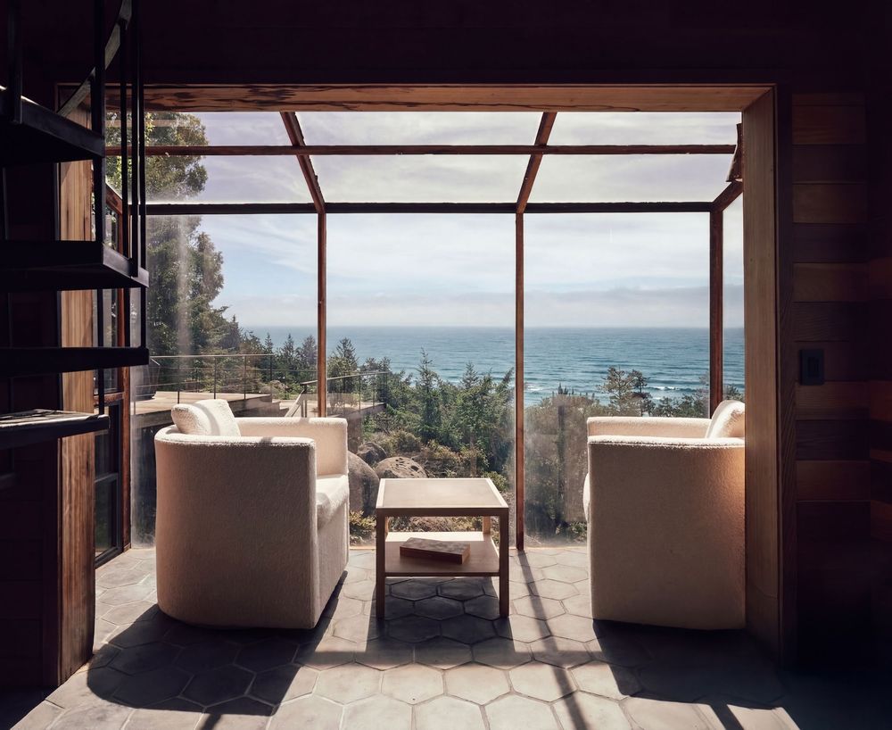 Modern living room with white chairs and ocean view through large glass windows overlooking coastal trees.