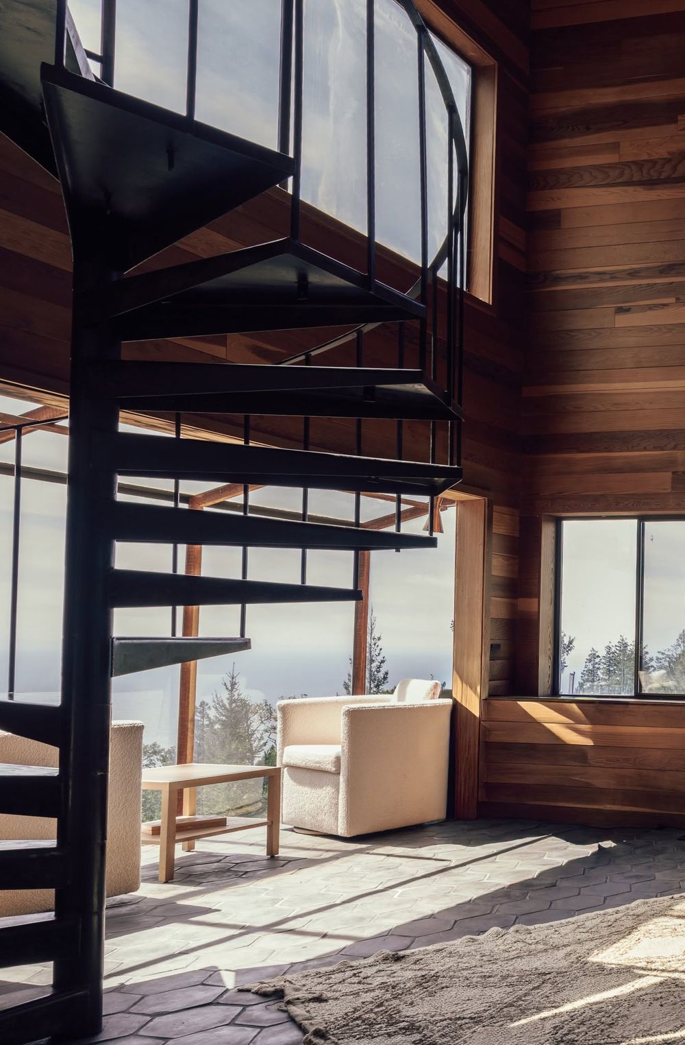 Black spiral staircase in a modern wood-paneled cabin with large windows and a white armchair overlooking a scenic view.
