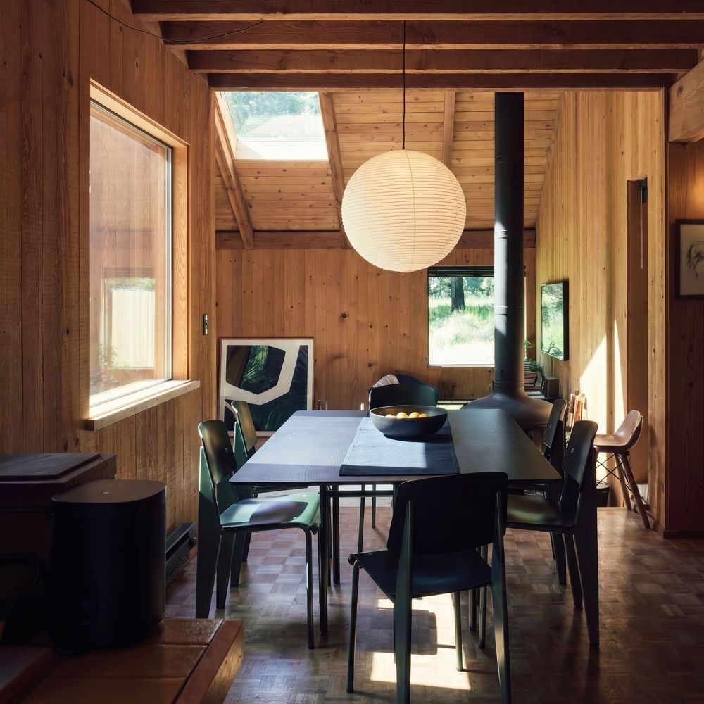 Rustic wood-paneled dining room with black table, globe pendant light, skylight, and wood-burning stove.
