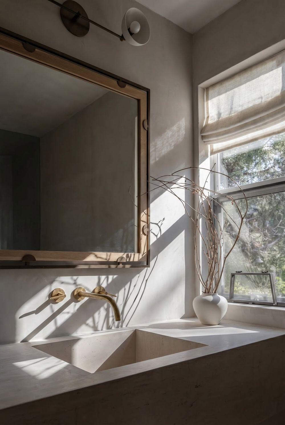 Minimalist bathroom with concrete sink, gold wall-mounted faucet, wood-framed mirror, and natural light from a linen Roman shade window.