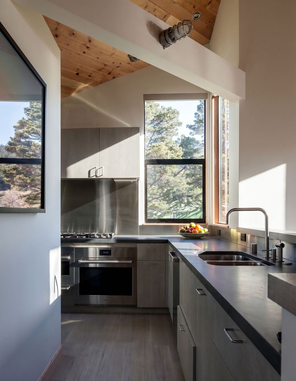 Modern kitchen with gray cabinets, stainless steel appliances, dark countertops, and large windows overlooking trees with wood ceiling.