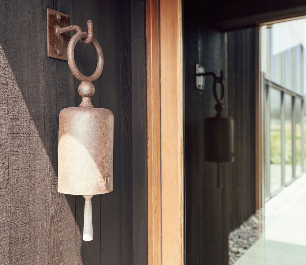 Rustic cast iron bell mounted on dark exterior wall beside wooden door, with reflection visible in glass.
