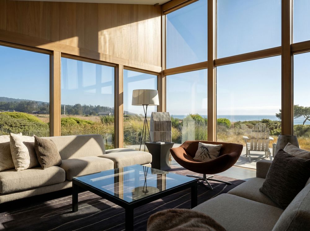 Bright living room with floor-to-ceiling glass walls, neutral sofas, and panoramic coastal meadow views under blue sky.