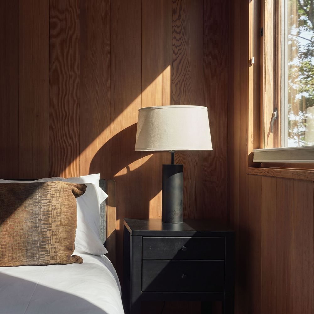 Cozy bedroom with dark wood paneling, black nightstand, table lamp, and white bedding near a sunlit window.