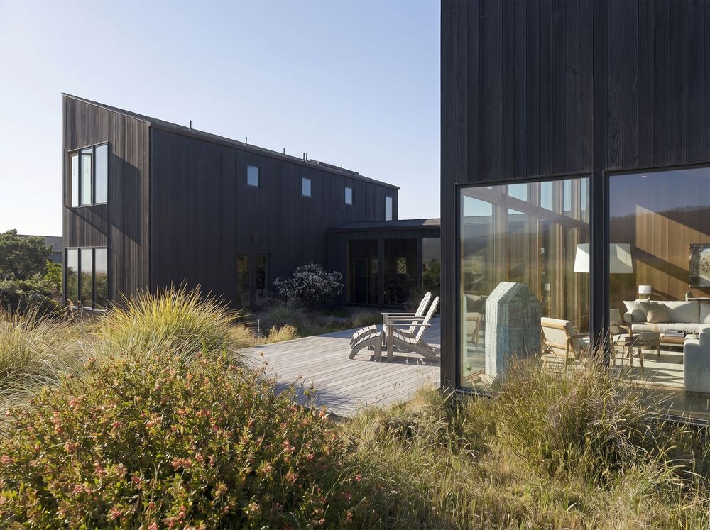 Dark timber clad coastal home with wooden deck, outdoor chairs, and native grasses under clear blue sky.