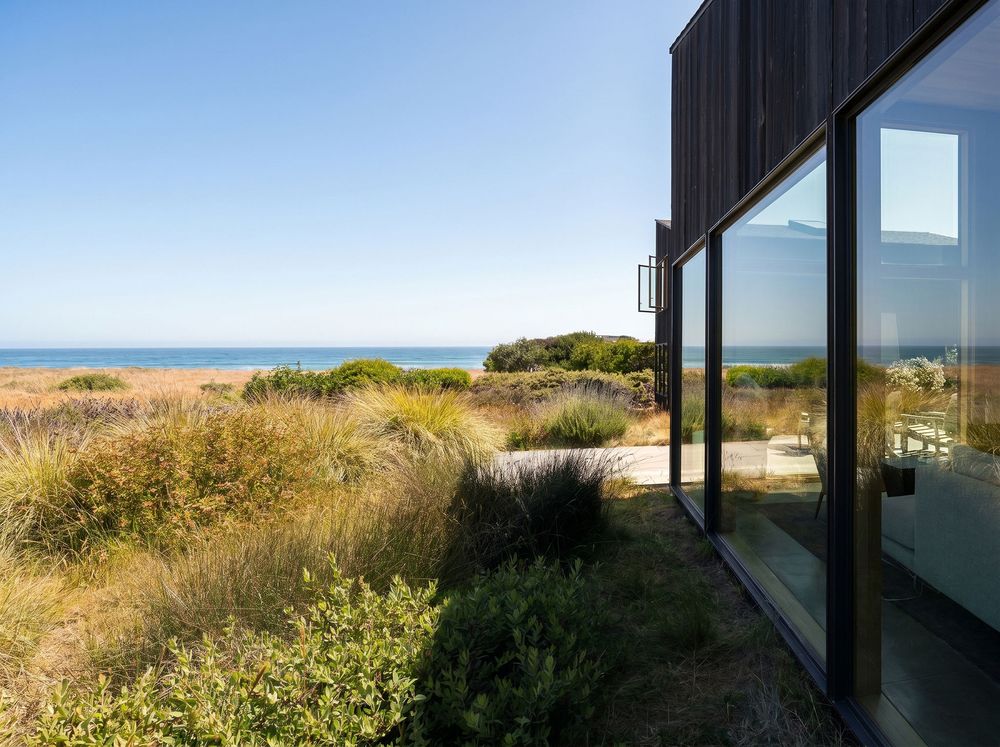 Modern dark-clad beach house with floor-to-ceiling glass windows overlooking coastal dunes and blue ocean under clear sky.