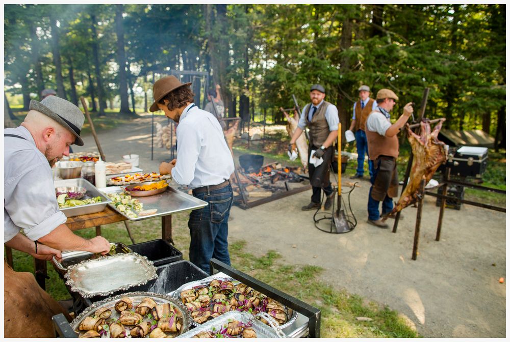 Outdoor barbecue event with chefs grilling food while guests gather around in a wooded setting.