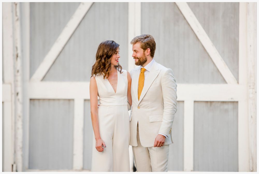 Couple in elegant white attire standing against rustic barn doors.
