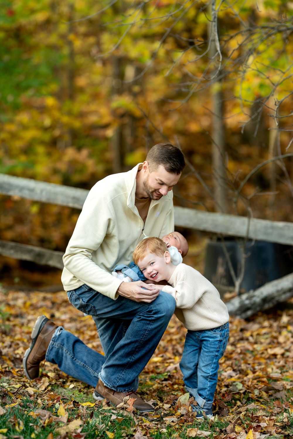 A laughing father kneels in fall leaves as his red-haired toddler son playfully leans against him holding the newborn.