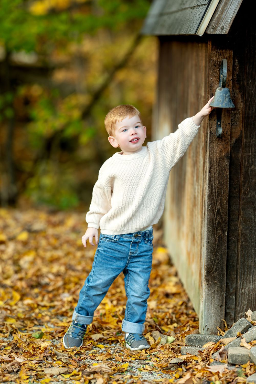 A cute red-haired toddler boy in a white sweater rings a bell on a rustic wooden structure in autumn leaves.