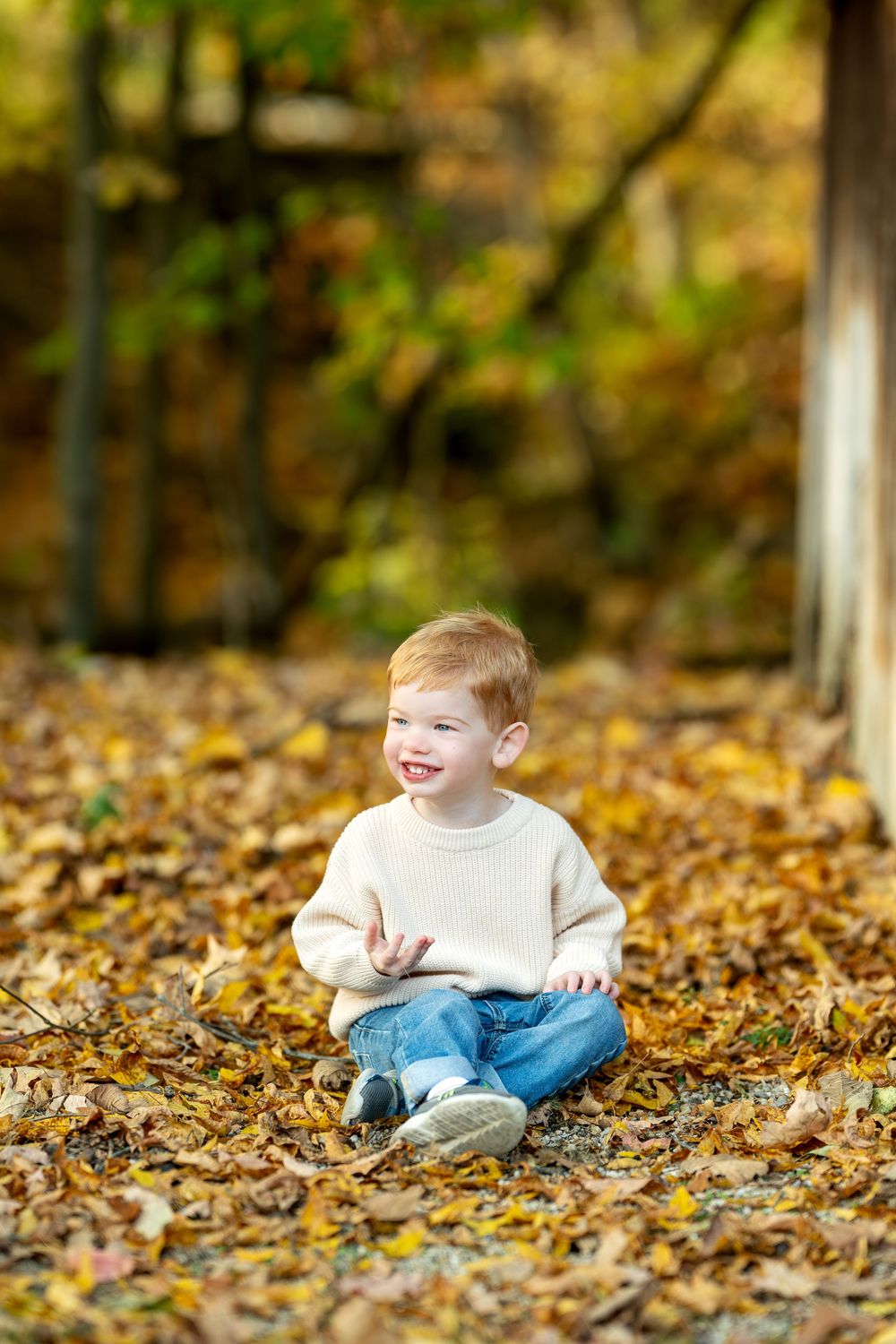 An adorable red-haired toddler boy sits cross-legged smiling in a pile of colorful autumn leaves outdoors.