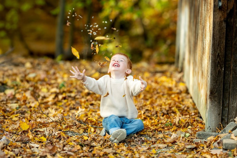 Happy toddler tossing golden fall leaves into the air outdoors.