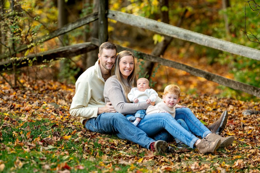 Happy family of four sitting together in colorful autumn leaves near wooden fence.