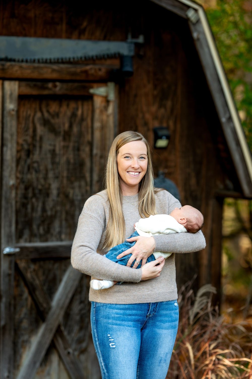 Smiling blonde mother holding sleeping newborn baby in front of rustic wooden barn.