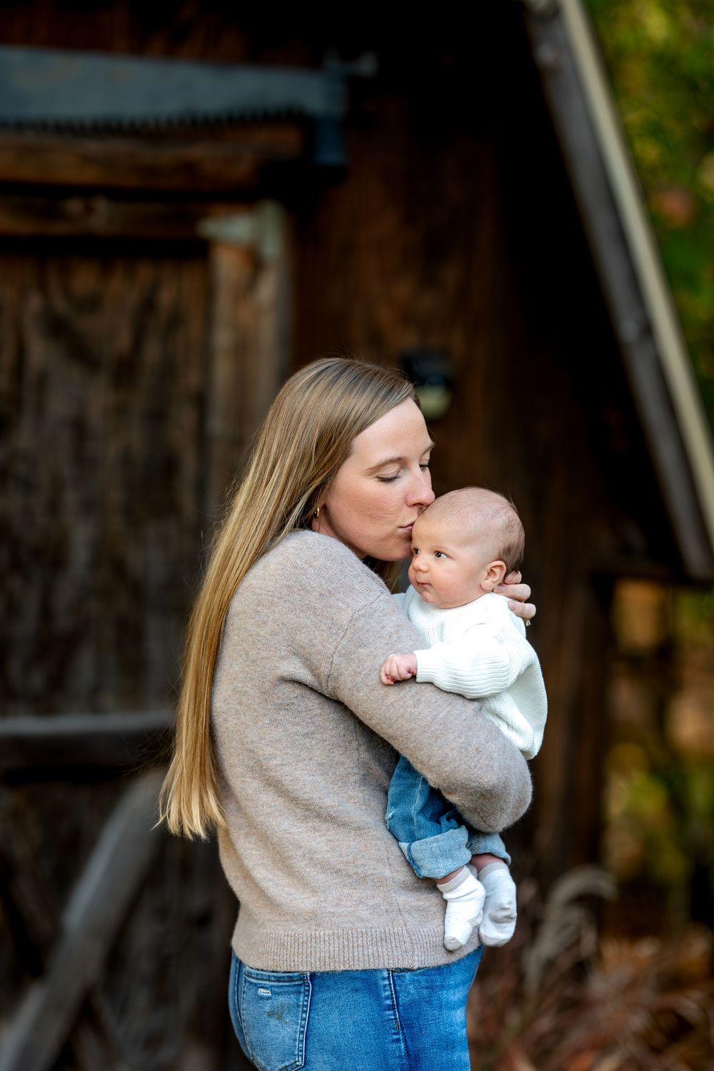 Loving mother kissing newborn baby while holding them near a rustic wooden barn.