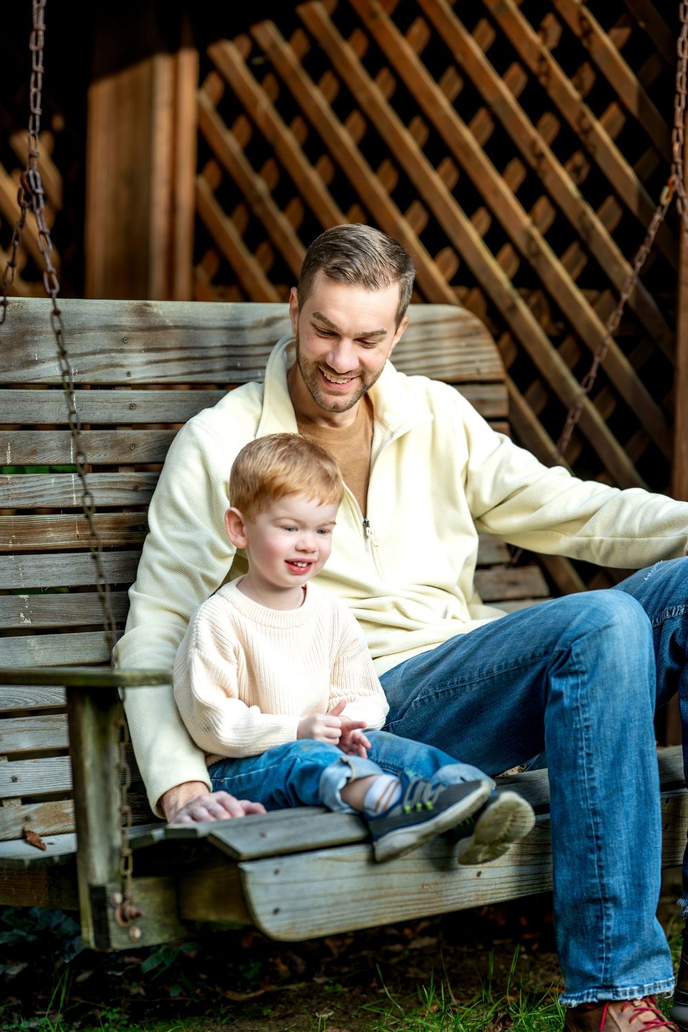 Father and toddler son sitting together on wooden porch swing, smiling outdoors.
