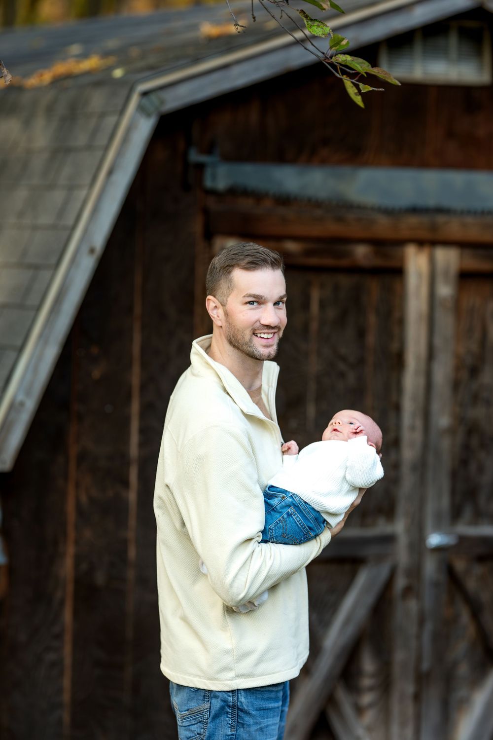 Happy father holding sleeping newborn baby outdoors near rustic wooden barn in fall.