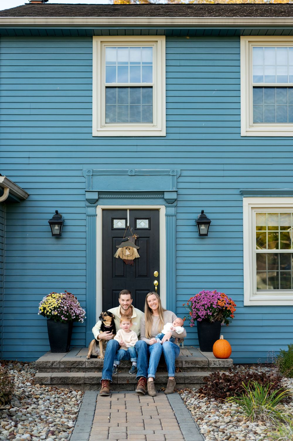 Family with two children and dog sitting on steps of blue colonial home in fall.