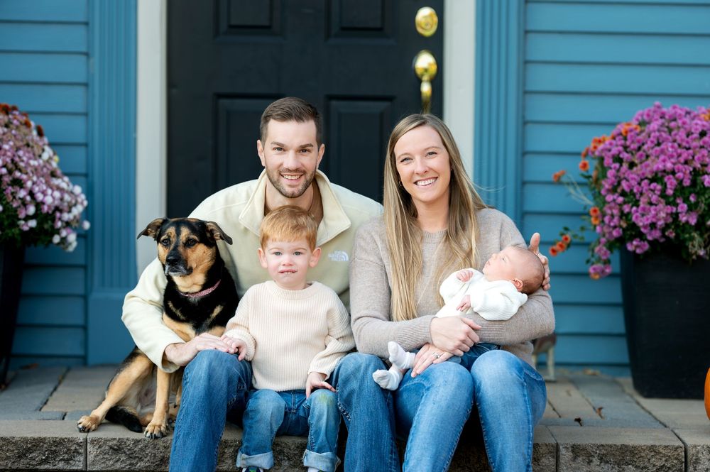 Family portrait with parents, toddler, newborn baby, and dog on blue house steps.