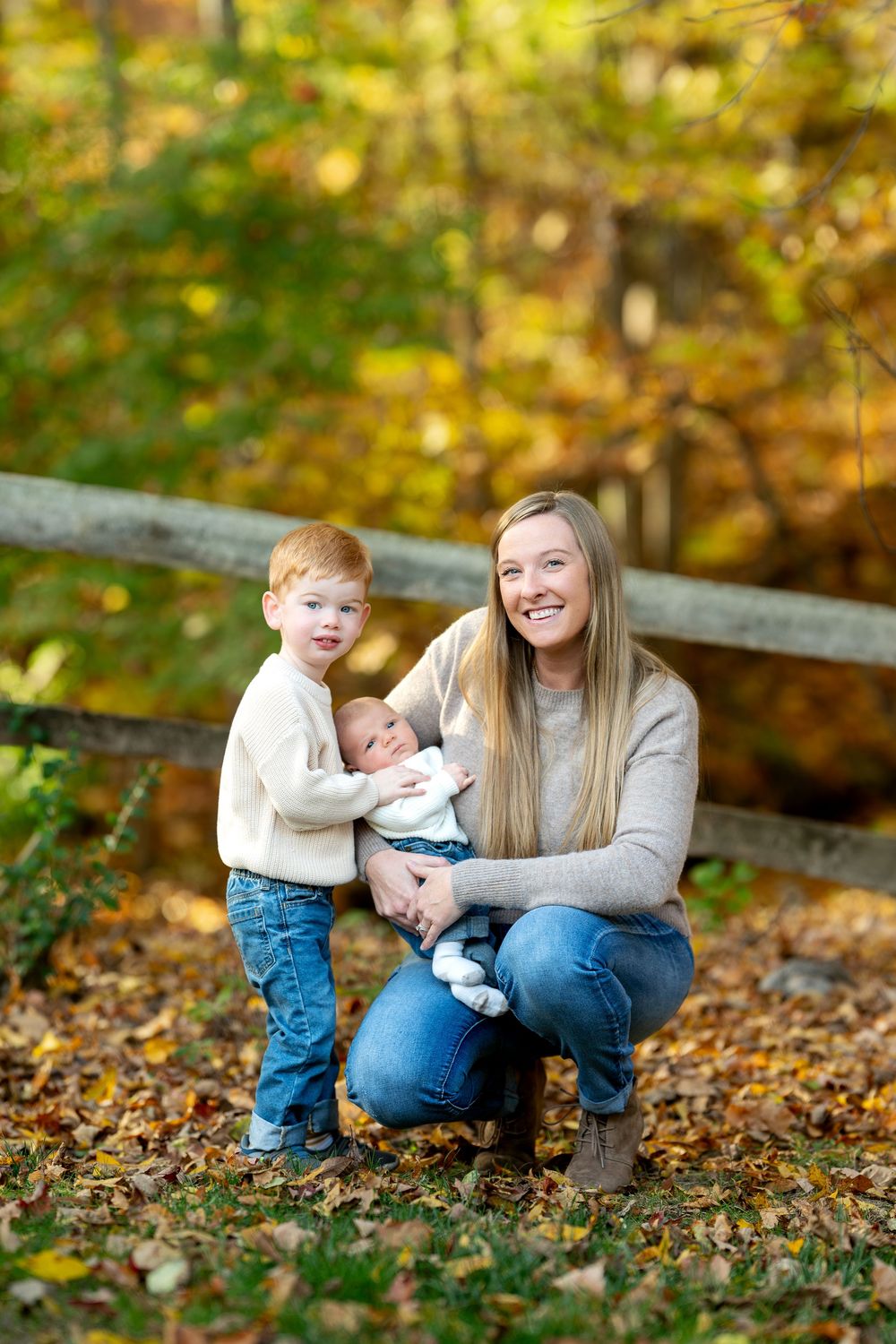 A smiling mother crouches with her toddler son and newborn baby amid fall foliage in an autumn park setting.