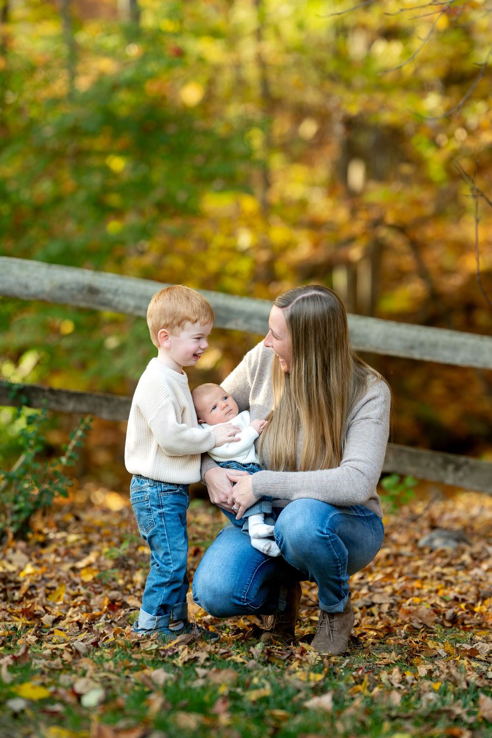 A mother kneeling in autumn leaves shares a tender moment with her laughing toddler and newborn baby outdoors.
