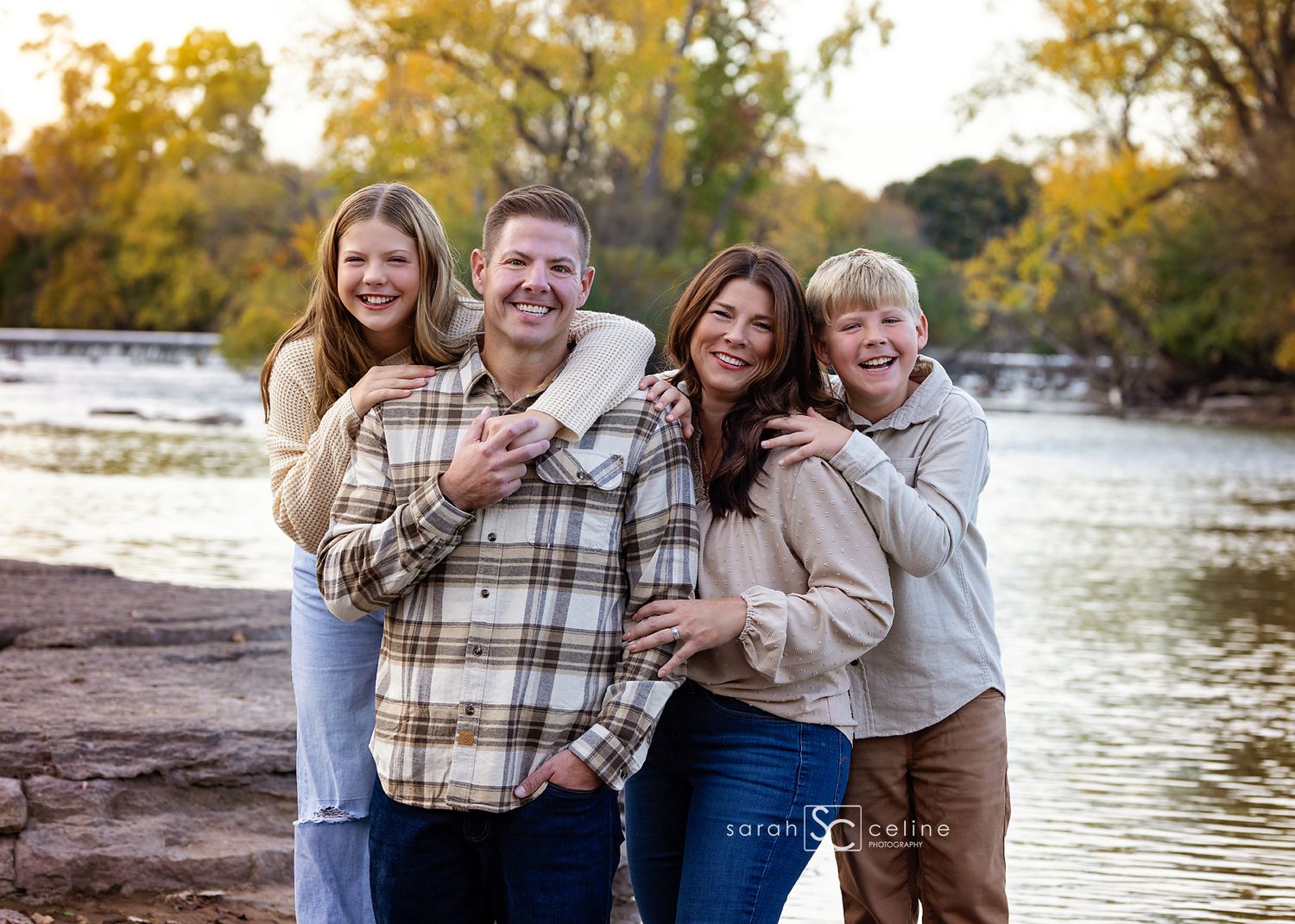 A group poses together outdoors by a river during autumn with golden trees and rocky shore in the background.