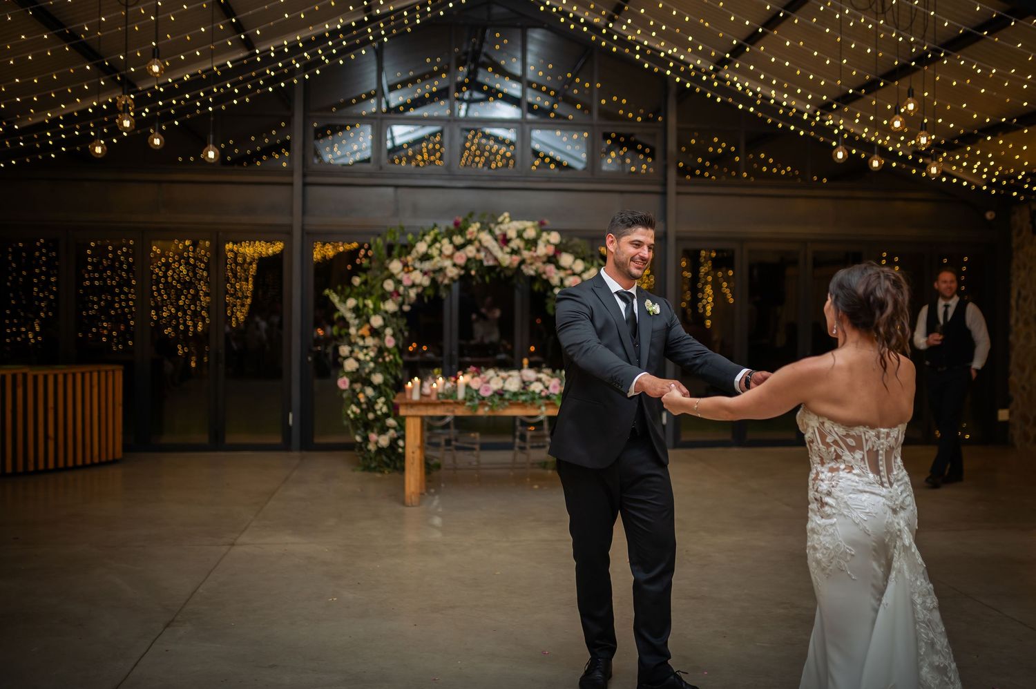 A couple dances together on a wooden floor under twinkling string lights at an indoor wedding reception.