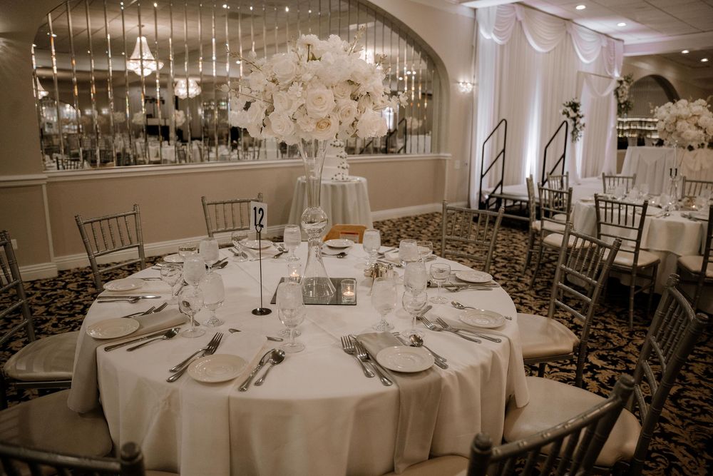 Elegant wedding reception table with white linens, silver chiavari chairs, and tall white floral centerpiece in ballroom setting.