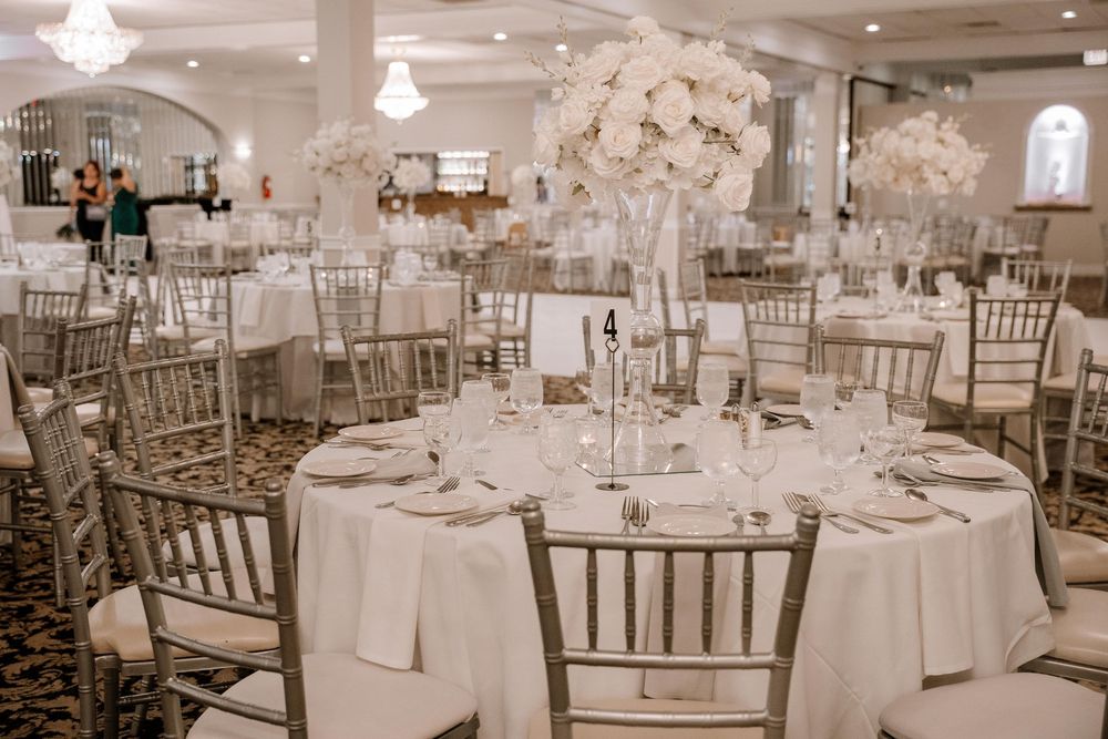 Elegant wedding reception hall with white decor, crystal chandeliers, and chiavari chairs arranged around round tables.