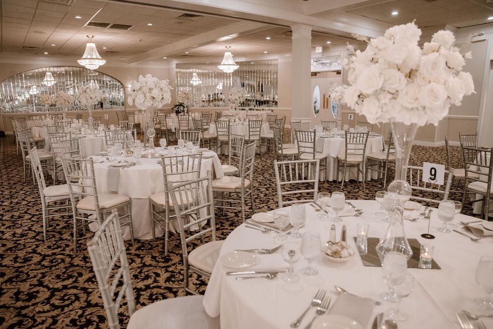Wedding reception venue with white chiavari chairs and elegant table settings under crystal chandeliers.