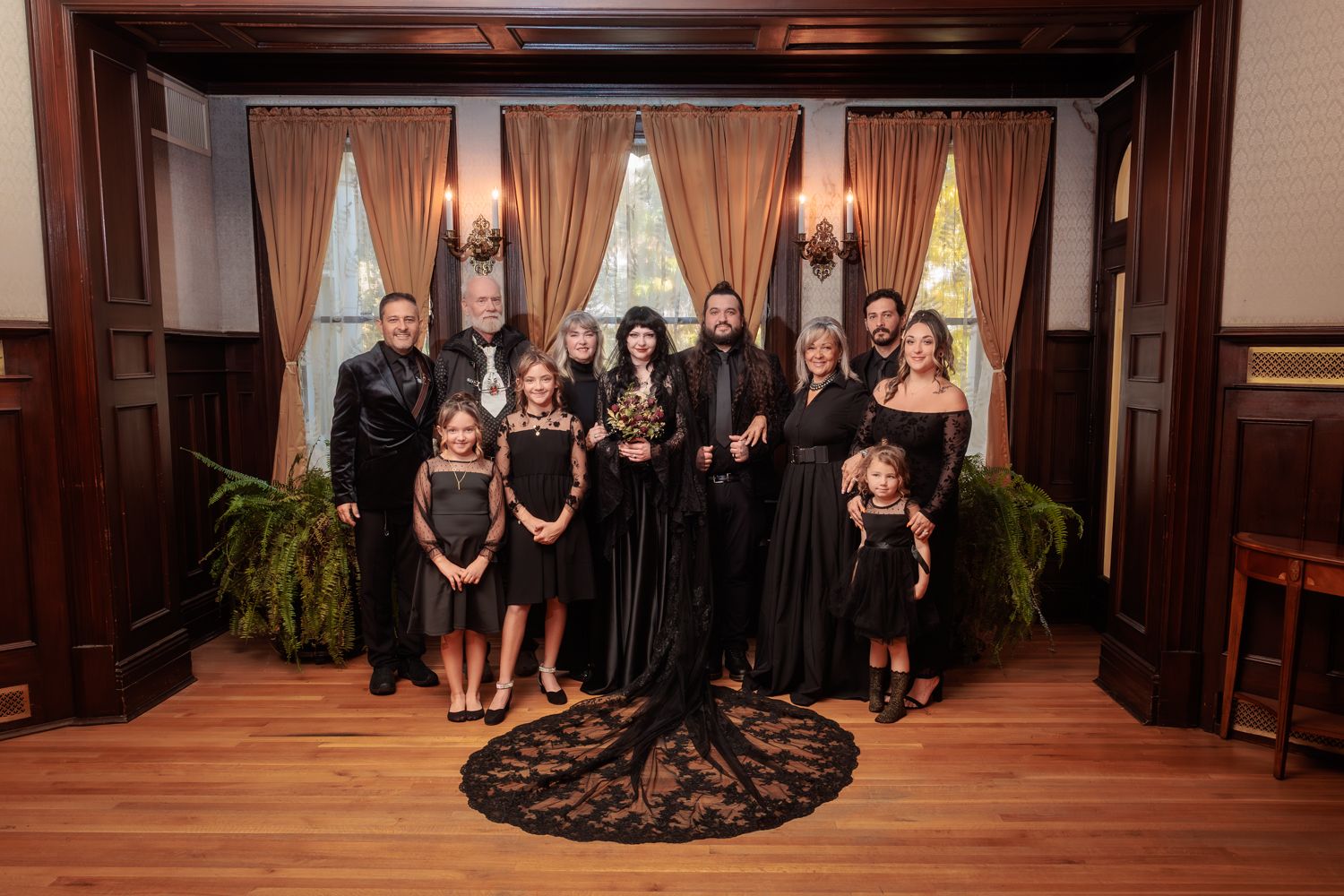 Wedding party in black attire poses formally in a grand wood-paneled room.