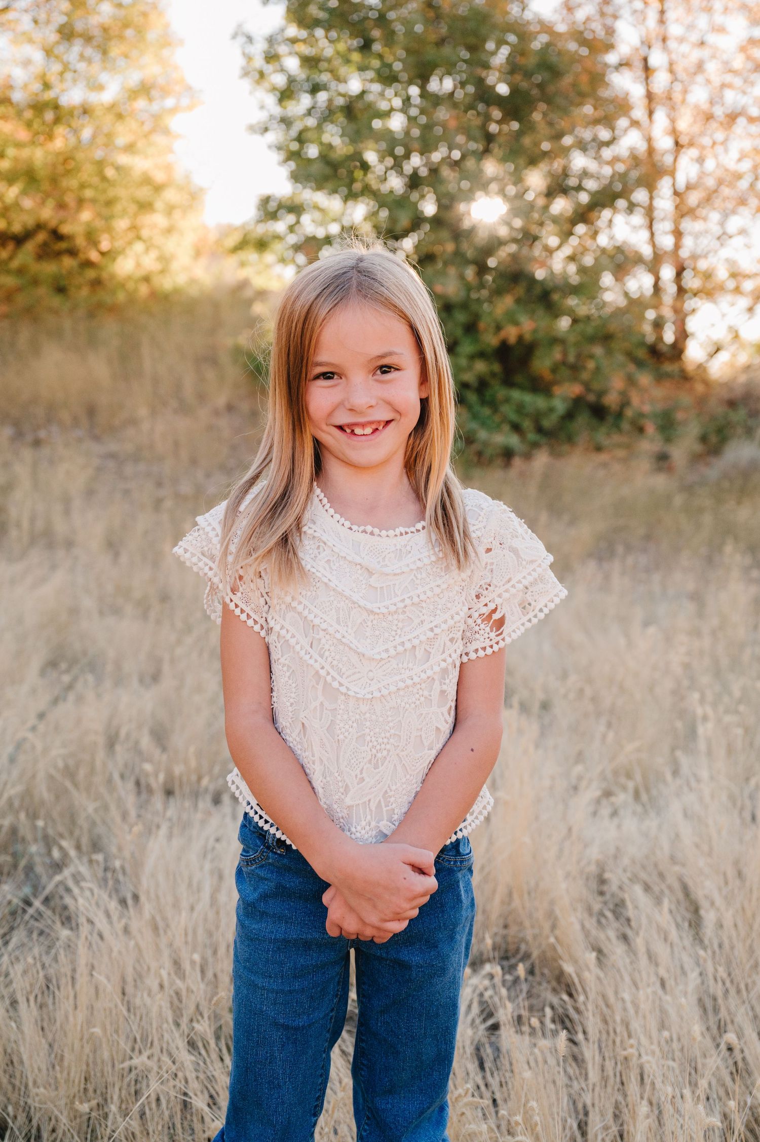 A young child in a white lace top and jeans poses playfully in a field of tall golden grass at sunset.