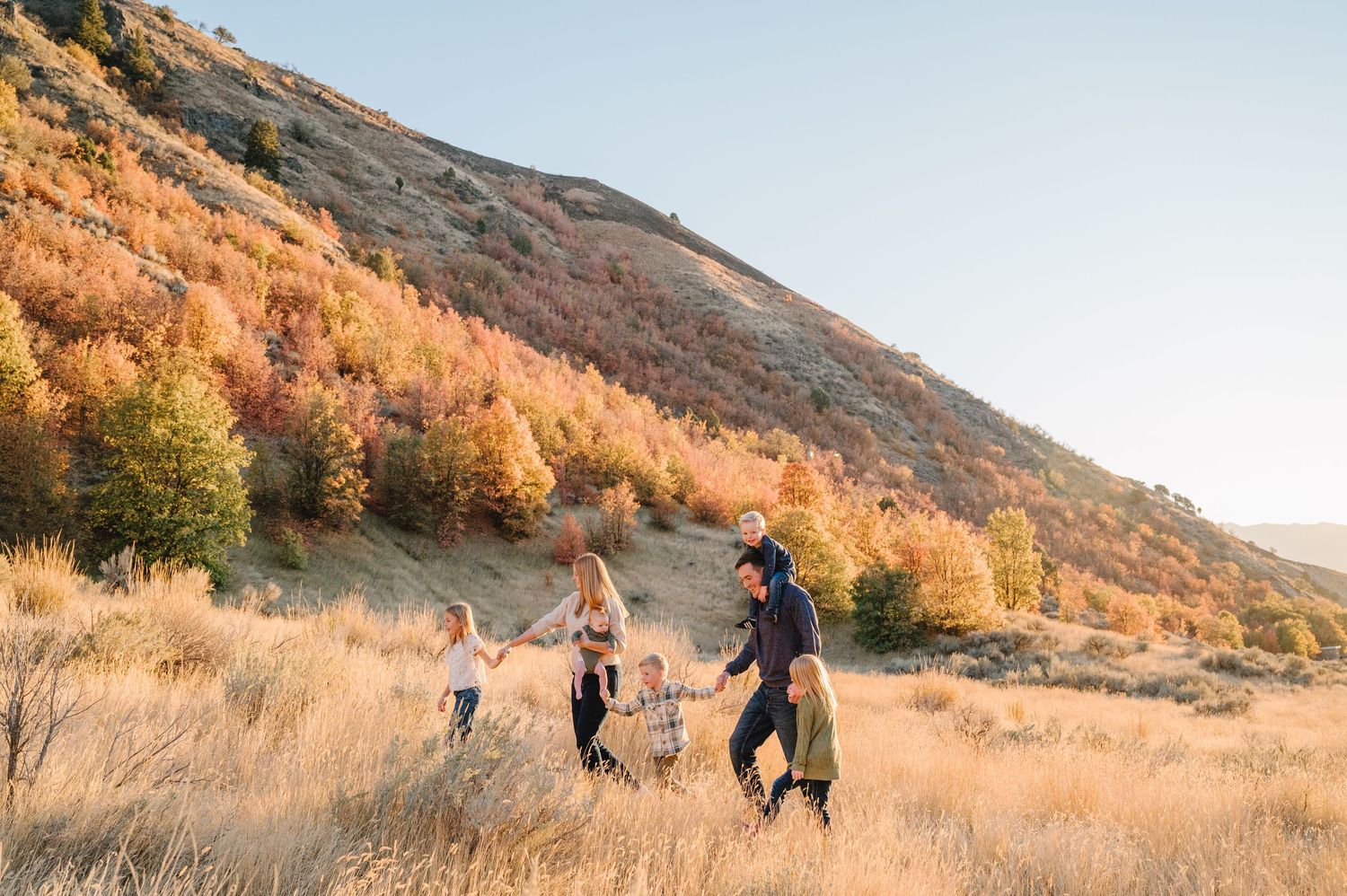A group of people walk along a scenic mountain trail at sunset with golden light illuminating the hillside.