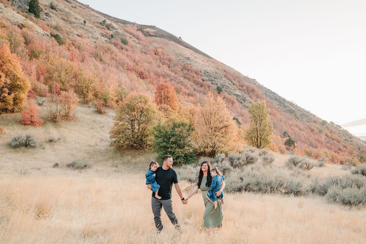Two hikers walk through a golden autumn meadow with colorful fall foliage on mountainside in the background.