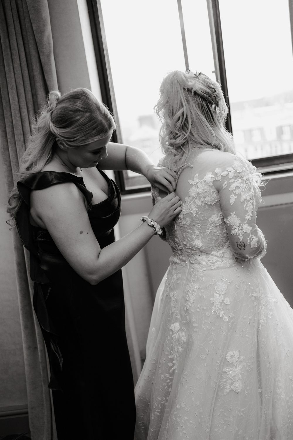 A tender moment captured in black and white as final adjustments are made to a beautiful lace wedding dress by a window.