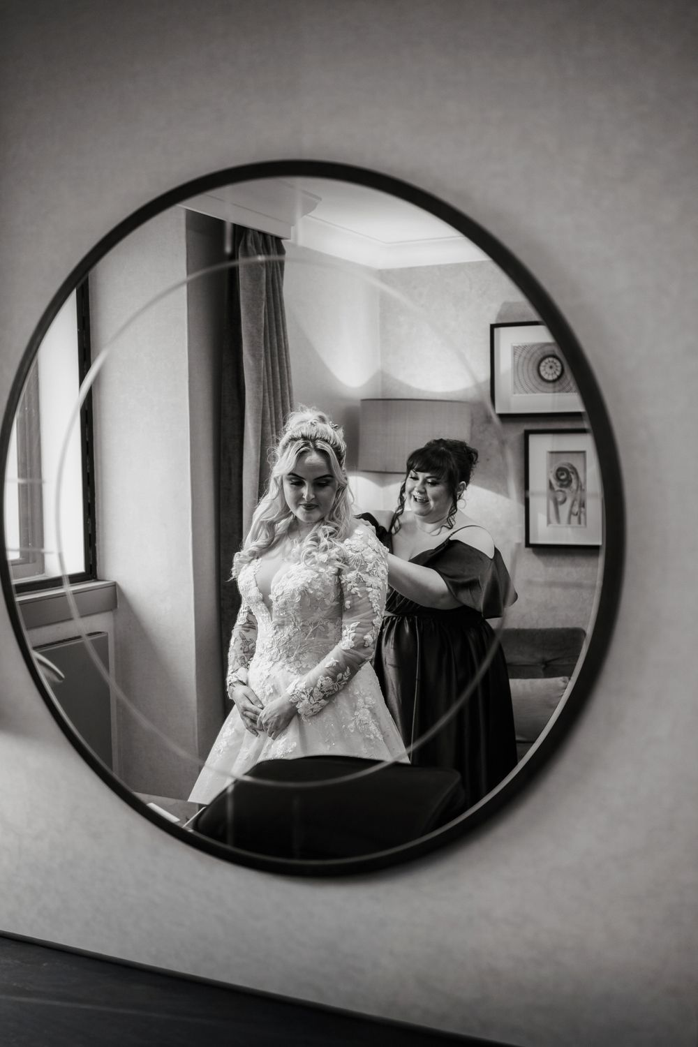 A circular mirror reflection shows the final touches being made to a bride's elegant long-sleeved lace gown.