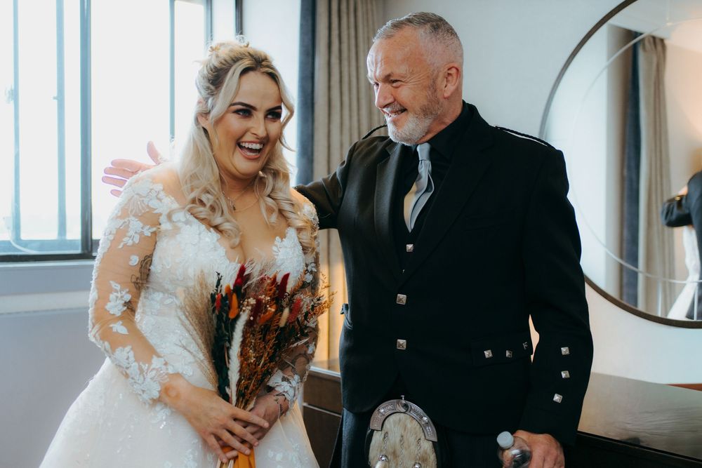A joyful bride in a lace wedding dress laughs while holding a bouquet next to someone wearing traditional Scottish attire.