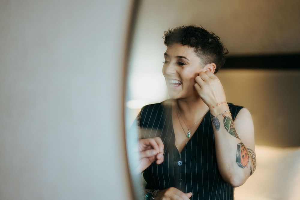 A person wearing a pinstripe vest smiles while touching their face in a softly lit indoor setting.