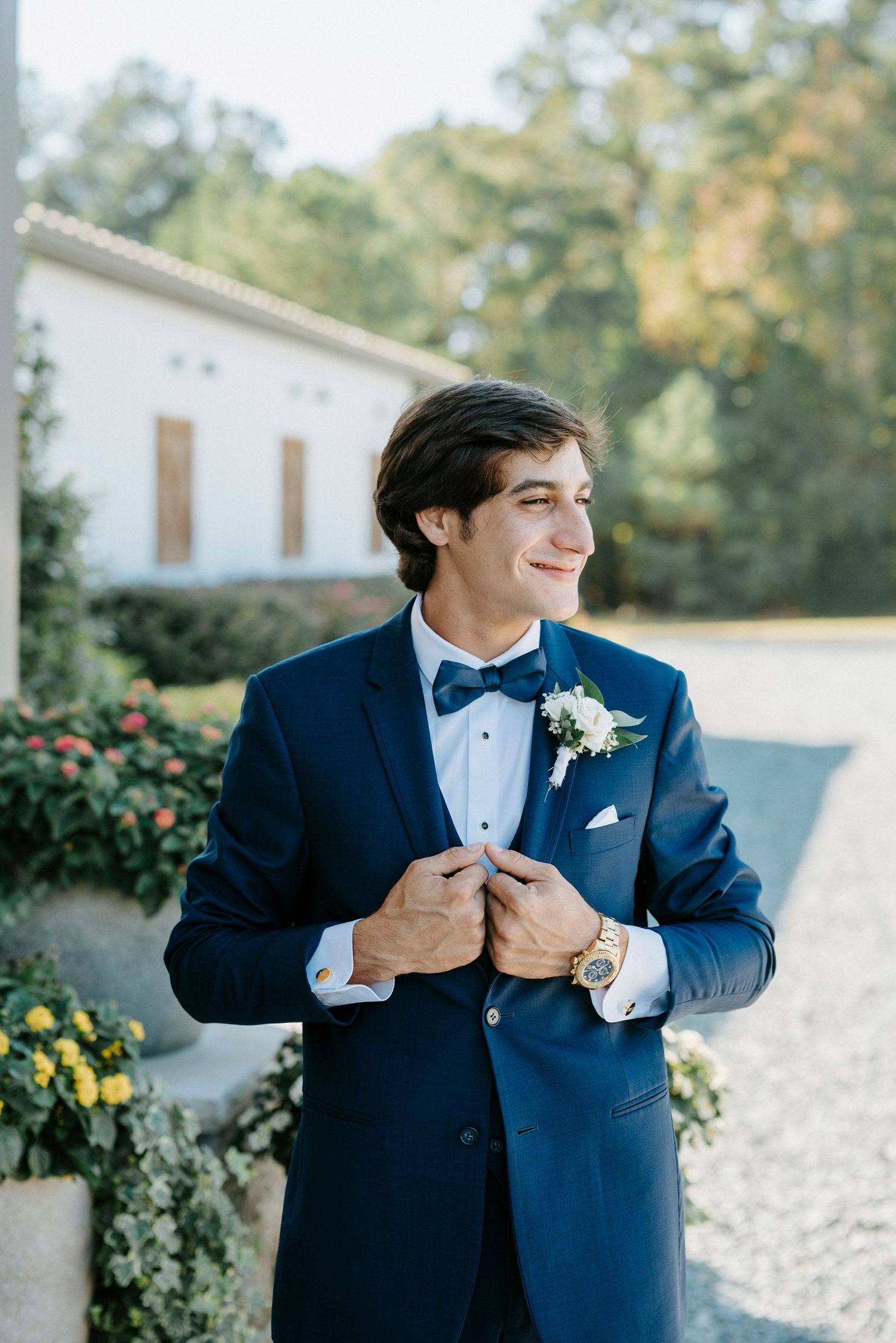 A groom in a navy blue tuxedo with bow tie and boutonniere smiles while adjusting his jacket outdoors.